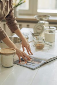 A woman in a cozy kitchen flipping through a cookbook while gathering ingredients for baking.